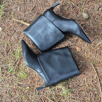 A pair of black leather ankle boots with pointed toes and block heels, placed on a bed of pine needles and dry grass outdoors.