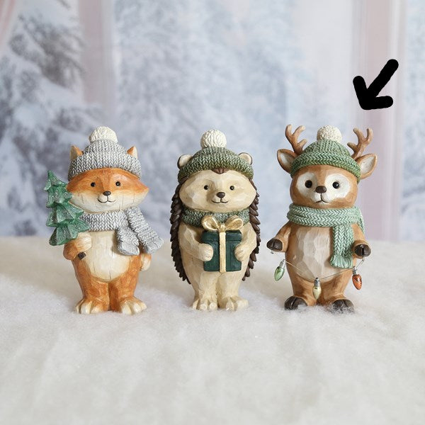 Three small winter-themed animal figurines standing side by side on a white surface against a soft snowy backdrop. The left figure is a fox in a gray knit hat and scarf holding a green tree, the center figure is a hedgehog in a green knit hat and scarf holding a gift box, and the right figure is a deer in a green knit hat and scarf with antlers.