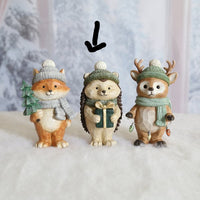 Three small winter-themed animal figurines standing side by side on a white surface against a soft snowy backdrop. The left figure is a fox in a gray knit hat and scarf holding a green tree, the center figure is a hedgehog in a green knit hat and scarf holding a gift box, and the right figure is a deer in a green knit hat and scarf with antlers.