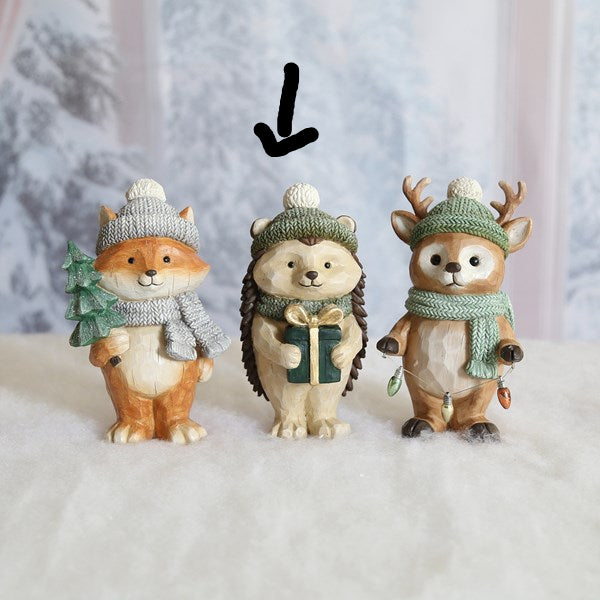 Three small winter-themed animal figurines standing side by side on a white surface against a soft snowy backdrop. The left figure is a fox in a gray knit hat and scarf holding a green tree, the center figure is a hedgehog in a green knit hat and scarf holding a gift box, and the right figure is a deer in a green knit hat and scarf with antlers.