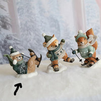 Three small animal figurines dressed in winter clothing posed on a snowy surface with a blurred winter forest backdrop. The figures appear to be a bear, fox, and squirrel wearing knit hats, sweaters, gloves, and skis.