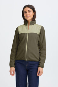 A woman modeling a color-blocked fleece jacket with an olive green body and light sage green yoke, paired with dark blue jeans, against a light gray studio background.