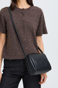 A person wearing a brown short-sleeve button-front top and dark jeans, carrying a black woven crossbody bag with a black strap against a light gray background.