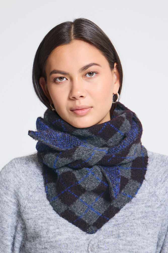 A woman wearing a thick, plaid infinity scarf in shades of navy blue, gray, and black, paired with a light gray knit sweater against a plain light gray background.