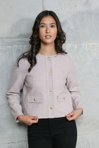 Woman wearing a light pink jacket with gold buttons against a textured gray wall.