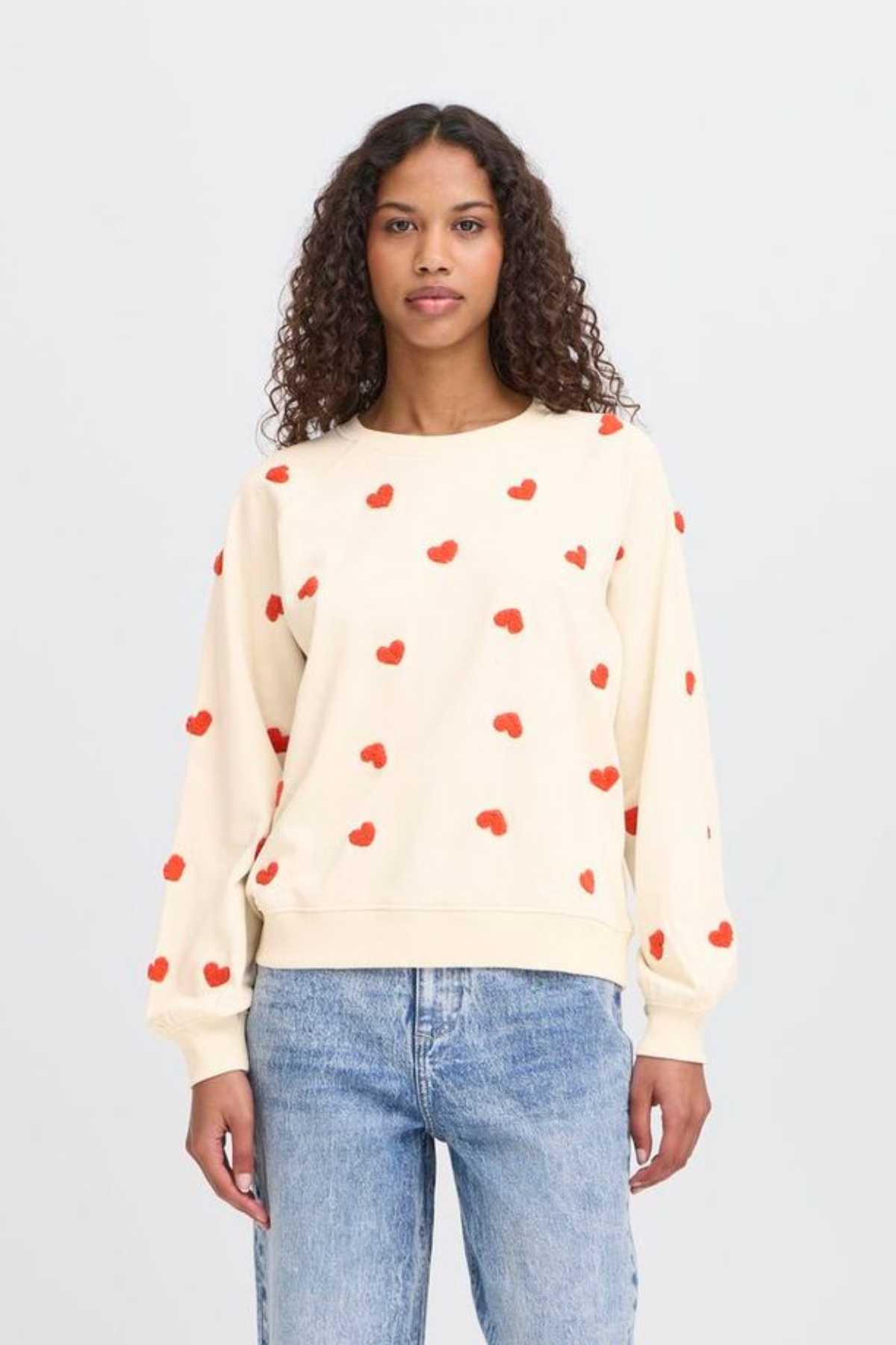 Woman wearing a cream sweater with red heart patterns and blue jeans on a light gray background