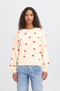 Woman wearing a cream sweater with red heart patterns and blue jeans on a light gray background