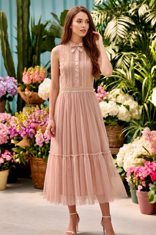 A woman models a sleeveless blush pink midi dress with a lace bodice, high neck, and tiered tulle skirt, set against a lush garden background with potted plants and flowers.